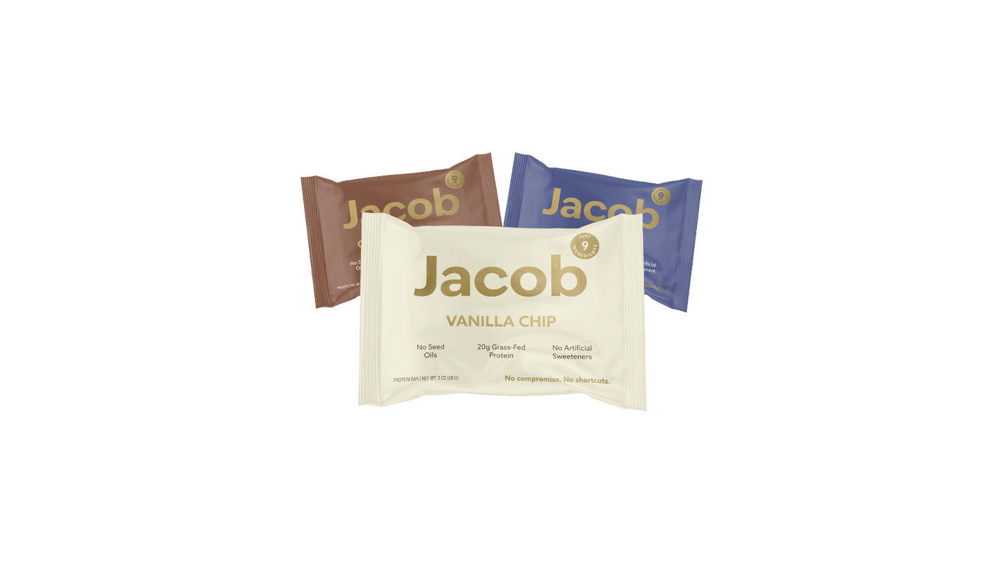 Three Jacob Bar snack packages are displayed. The front package is Vanilla Chip in beige, while behind it are two other packages: one in brown and another in dark blue, with indistinct flavor labels. Made with 100% Organic Cacao, these bars contain no seed oils, soy, or artificial sweeteners.