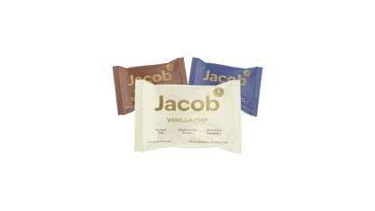 Three Jacob Bar snack packages are displayed. The front package is Vanilla Chip in beige, while behind it are two other packages: one in brown and another in dark blue, with indistinct flavor labels. Made with 100% Organic Cacao, these bars contain no seed oils, soy, or artificial sweeteners.