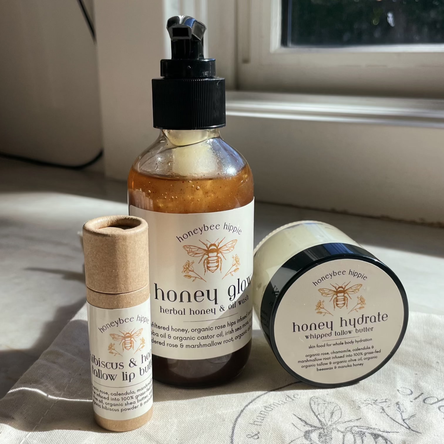 Skincare products shine in natural light: "The Honey Bundle" by Honeybee Hippie, featuring a honey glow body pump bottle, creamy whipped tallow butter in a jar, and a lip butter tube.