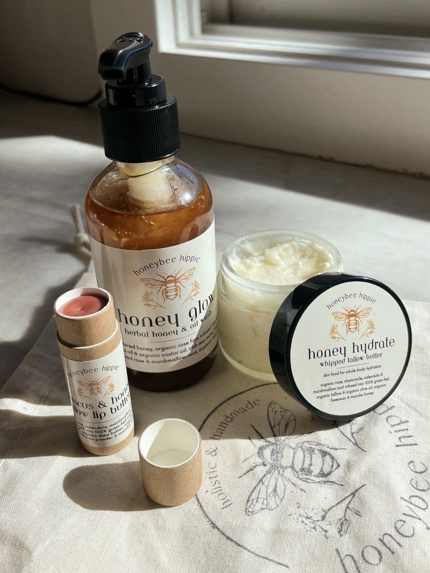 A collection of skincare products, "the honey bundle" by Honeybee Hippie, is displayed on a sunlit surface. It includes a pump bottle with anti-inflammatory "honey glow," whipped body butter, and tinted lip balm. The labels feature a bee motif.