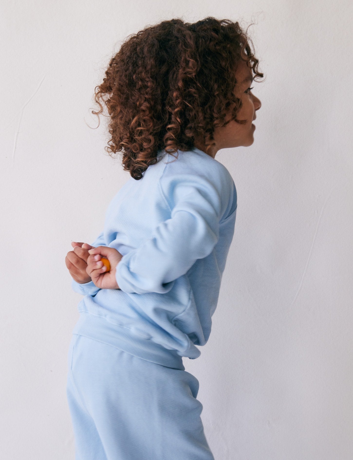 A young child with curly hair is standing sideways against a plain background. They are wearing matching light blue organic French terry cloth sweatshirt and pants from all the babies, with their hands clasped behind their back. The Lil' Classic Crewneck adds to the charm as the child appears to be smiling or laughing.