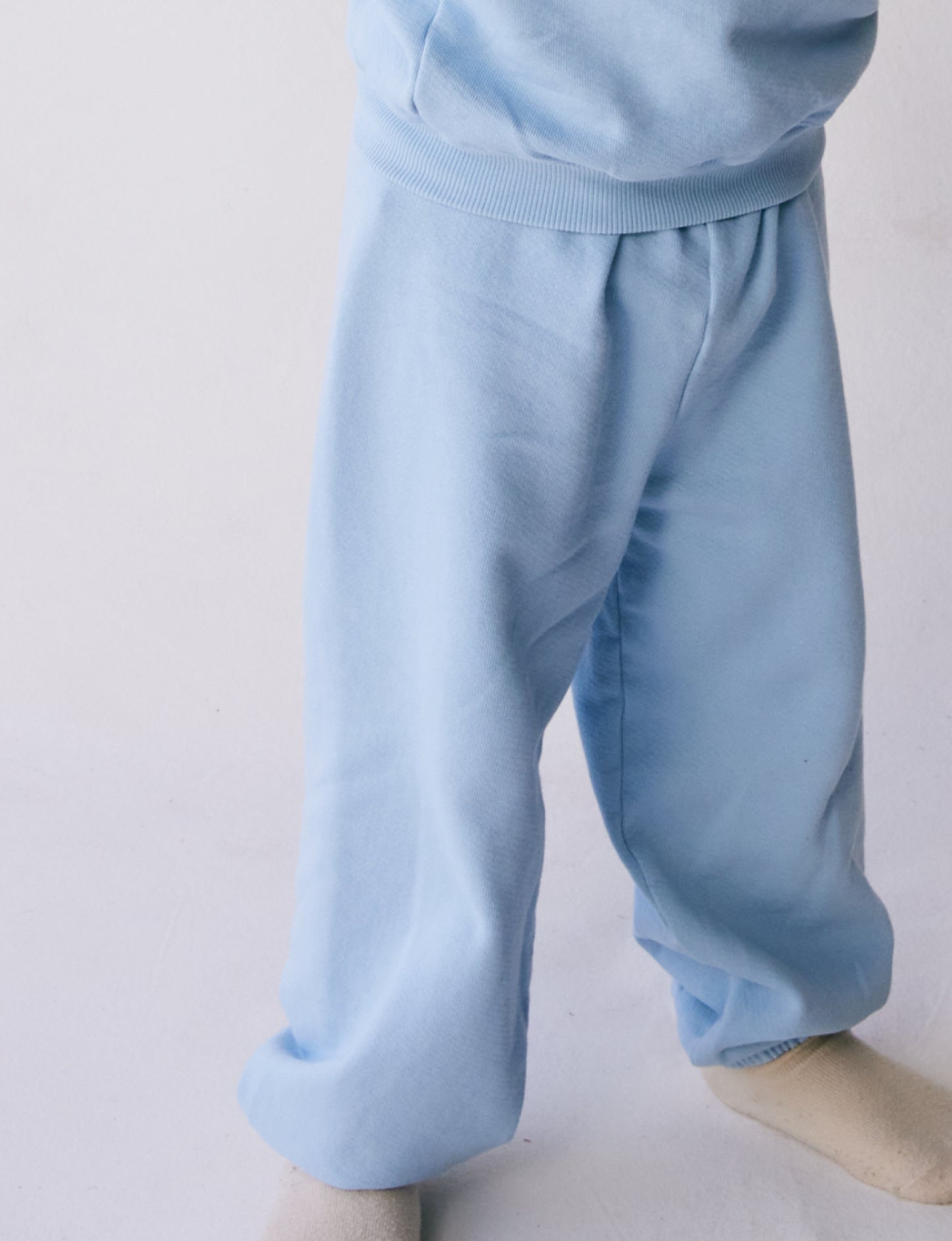 A child wearing **The Lil' Classic Sweatpant by all the babies**, crafted from organic French terry cloth, along with off-white socks stands against a plain background. The image focuses on the lower part of the child's body, showcasing the cozy outfit in detail.