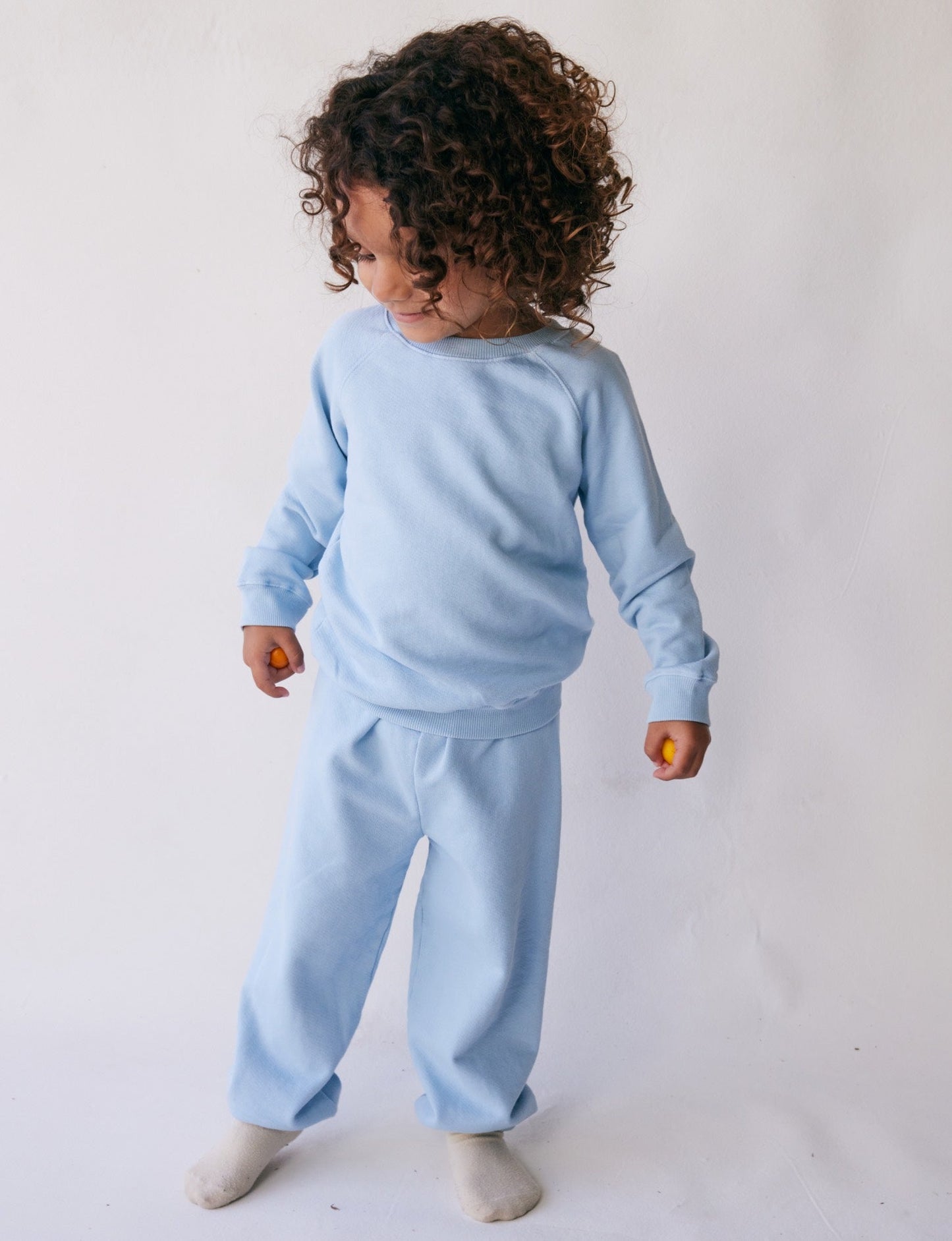 A young child with curly hair stands against a plain white background. The child is wearing The Lil' Classic Crewneck by all the babies, crafted from organic French terry cloth, paired with matching pants. They are looking down and to the side, holding a small object in their hand and wearing white socks.