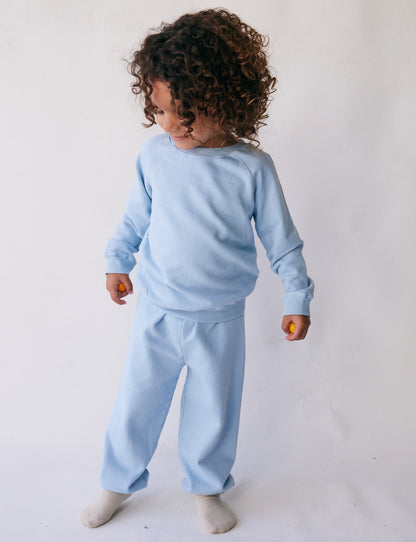 A young child with curly hair stands against a plain white background, wearing a light blue long-sleeve shirt and The Lil' Classic Sweatpant from all the babies, made of organic French terry cloth. The child has white socks on and is looking down to the side, holding a small object in their left hand.