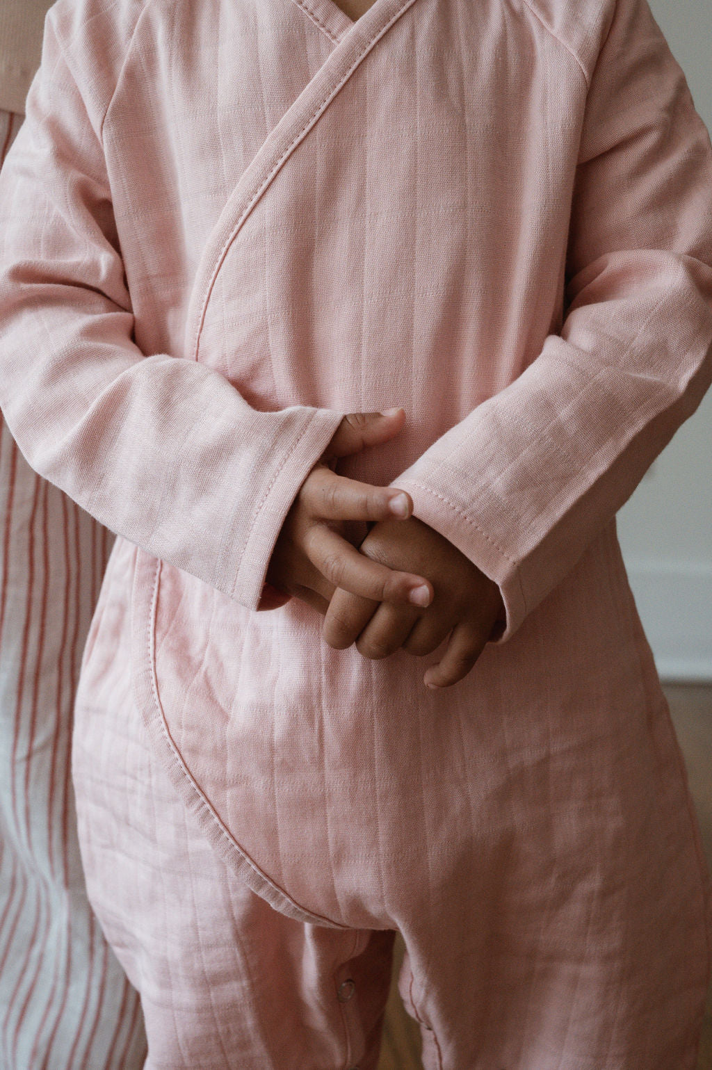 A young child wearing the Après Wrap in Desert Rose, a light pink, long-sleeved organic cotton double gauze wrap-style romper from cabane childrenswear, stands with their hands clasped together. The child is facing forward, and only their torso and part of their arms are visible in the image. Perfect for sensitive skin, the romper is ethically handmade.