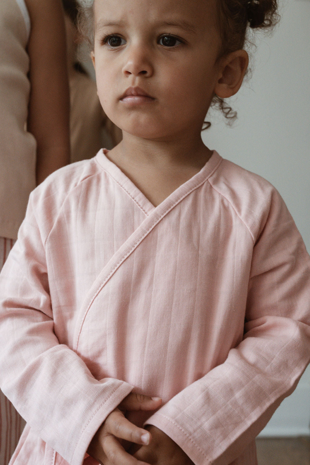 A young child with curly hair stands indoors, dressed in an ethically handmade Après Wrap in Desert Rose by cabane childrenswear. Looking slightly to the side with a serious expression, their hands are gently clasped in front of them. The background is softly lit and minimalistic.