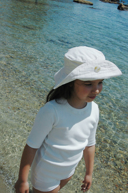 A child dressed in a white outfit and the "FRIENDS OF THE SUN" hat by cabane childrenswear stands on a shallow, clear beach with rocks in the background, gazing off to the side. The sunlight reflects off the water, creating a peaceful coastal scene, while the breathable summer material ensures comfort with every gentle breeze.