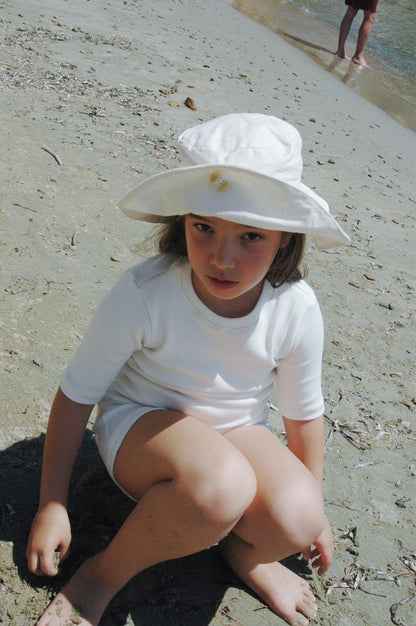 A young child, adorned in cabane childrenswear's classic "FRIENDS OF THE SUN" wide brim hat and white clothing made from breathable summer material, sits on the sandy beach with a serious expression. The ocean in the background and an adult's legs further down the shore add to the scene's serene atmosphere.