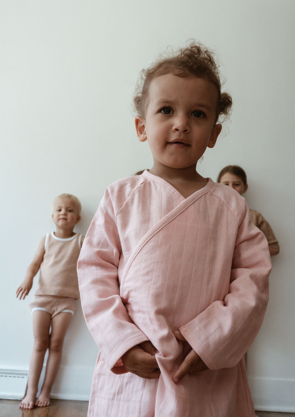 Three children stand against a plain white wall. The child in the foreground, dressed in the Après Wrap in Desert Rose from cabane childrenswear with a light pink long-sleeve top, exudes comfort for sensitive skin. The child on the left wears a beige sleeveless top and white shorts, while the one on the right sports a brown top. They all look toward the camera.