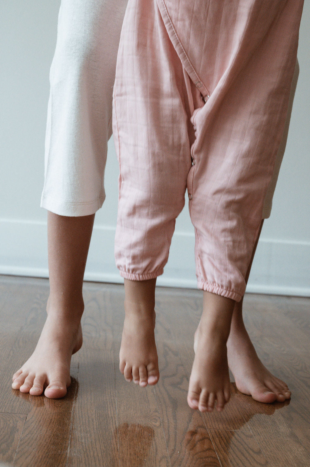 Two pairs of legs are shown, one belonging to an adult and the other to a child. The adult is wearing white pants, while the child is wearing pink Après Wrap in Desert Rose from cabane childrenswear, made from organically sourced cotton, perfect for sensitive skin. Both are barefoot and standing on a wooden floor.