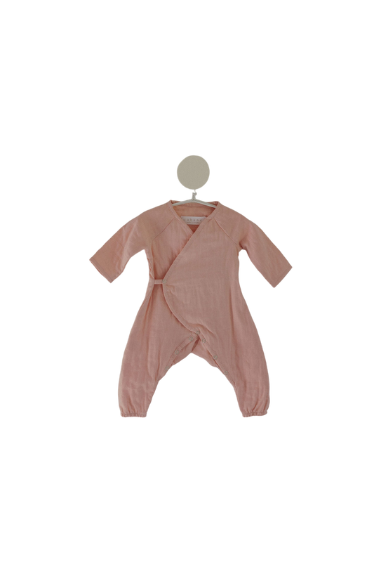 The Après Wrap in Desert Rose by cabane childrenswear is displayed on a white hanger. This ethically handmade, long-sleeved baby romper features a criss-cross front design and enclosed feet. Made from organic cotton double gauze wrap, it is soft and comfortable, perfect for infants with sensitive skin.