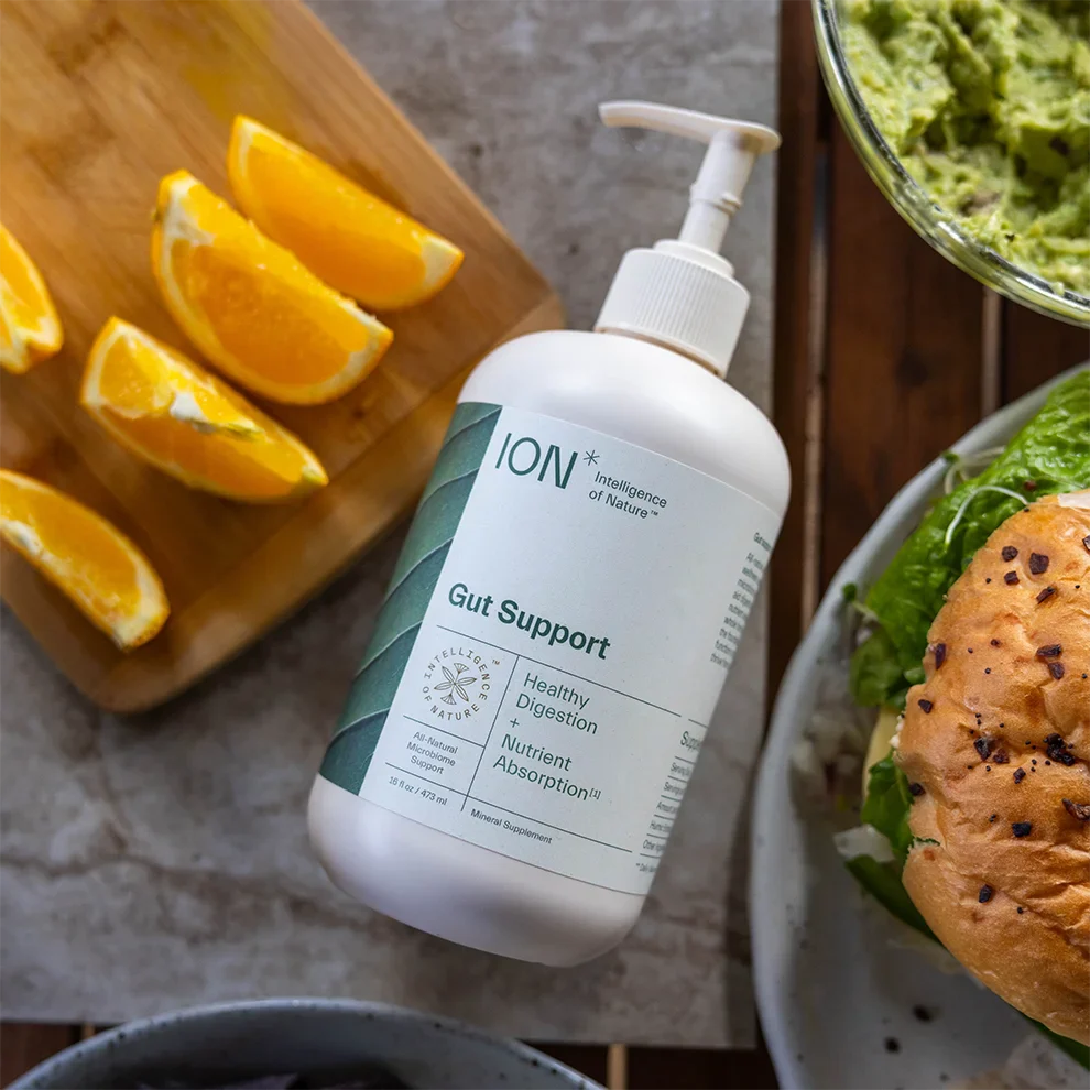 A bottle of ION* Gut Support stands on a table surrounded by food items, including orange wedges on a cutting board, a bowl of guacamole, and part of a sandwich with lettuce. This daily wellness supplement from ION* offers benefits such as healthy digestion, nutrient absorption, and microbiome support.