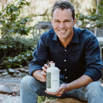 A person with short curly hair, wearing a dark blue shirt and jeans, is sitting outdoors on a stone. They are holding a bottle labeled "ION* Gut Support," a daily wellness supplement from ION* designed to support the microbiome, and smiling at the camera. The background features greenery and a blurred natural environment.