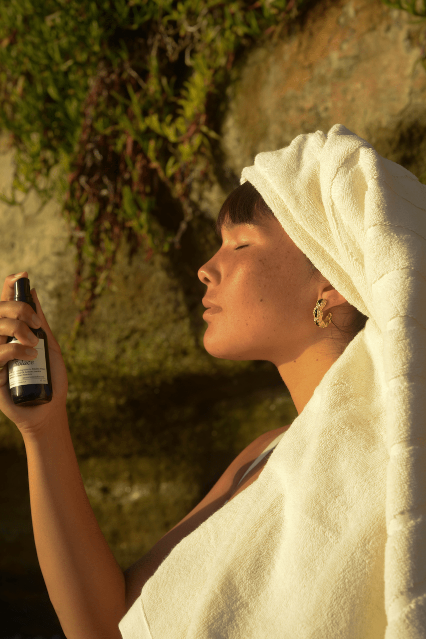 A person with a towel wrapped around their head is facing sideways under warm sunlight, eyes closed, and holding a bottle of Summer Solace Tallow's Jasmine Sambac (with Rose Ormus) Water-Based Perfume Mist near their face. They are standing against a backdrop of rocky terrain and greenery.