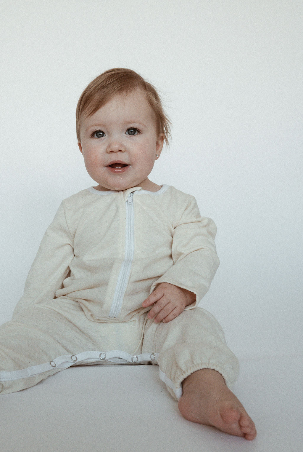 A smiling baby with light brown hair, dressed in THE ORB SUIT WAFFLE by cabane childrenswear, designed for easy diaper changes, sits against a plain white background. The baby has one hand resting on their lap and appears cheerful and happy.