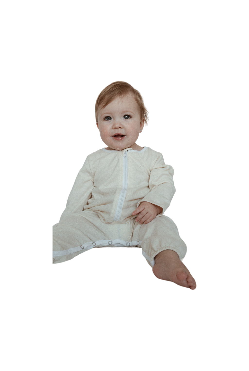 A baby with light brown hair sits on the floor wearing cabane childrenswear's THE ORB SUIT WAFFLE baby + kid, a light beige, full-body outfit featuring a zipper and snap buttons along the legs for easy diaper changes. Ethically handmade, this seasonless and unisex garment is perfect for any occasion. The baby looks ahead with a slight smile, legs extended with one slightly bent.