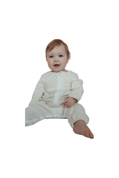 A baby with light brown hair sits on the floor wearing cabane childrenswear's THE ORB SUIT WAFFLE baby + kid, a light beige, full-body outfit featuring a zipper and snap buttons along the legs for easy diaper changes. Ethically handmade, this seasonless and unisex garment is perfect for any occasion. The baby looks ahead with a slight smile, legs extended with one slightly bent.