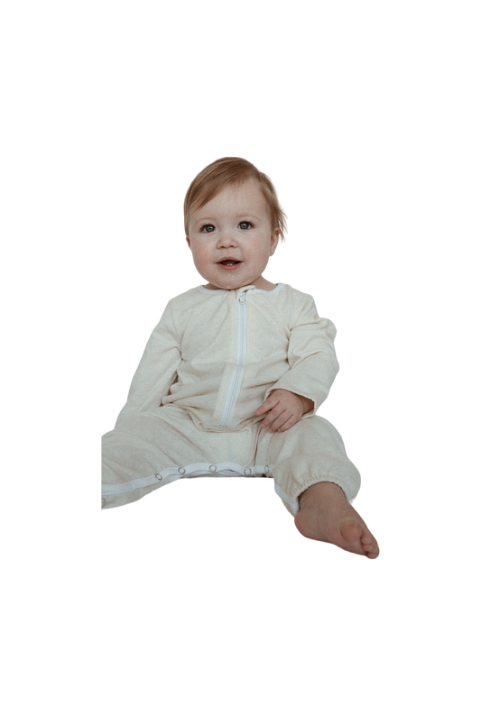 A happy baby with light brown hair sits upright, wearing a seasonless unisex outfit from cabane childrenswear. The ethically handmade white Orb Suit Waffle Baby + Kid features a front zipper and snap buttons on the legs, perfectly complementing the baby's relaxed posture with one leg bent and the other extended forward.