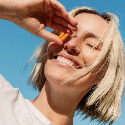 A smiling woman with blonde hair holds two Kinder Thoughts earplugs by THE FULLEST near her eye, their vibrant color reminiscent of mood-boosting turmeric. She stands against a clear blue sky, squinting slightly in the sunlight and appearing relaxed and happy.