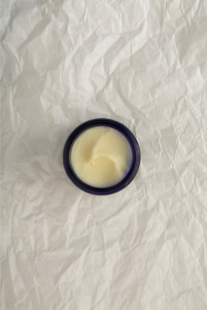 An open, dark blue jar of Lavendula Officinalis Balm - Regenerative Tallow™ from Summer Solace Tallow rests on a crumpled, white paper surface. The creamy, off-white lavender-scented balm features a smooth and slightly glossy texture that hints at the luxurious moisturizing experience it offers. The jar's circular shape casts a subtle shadow on the paper.