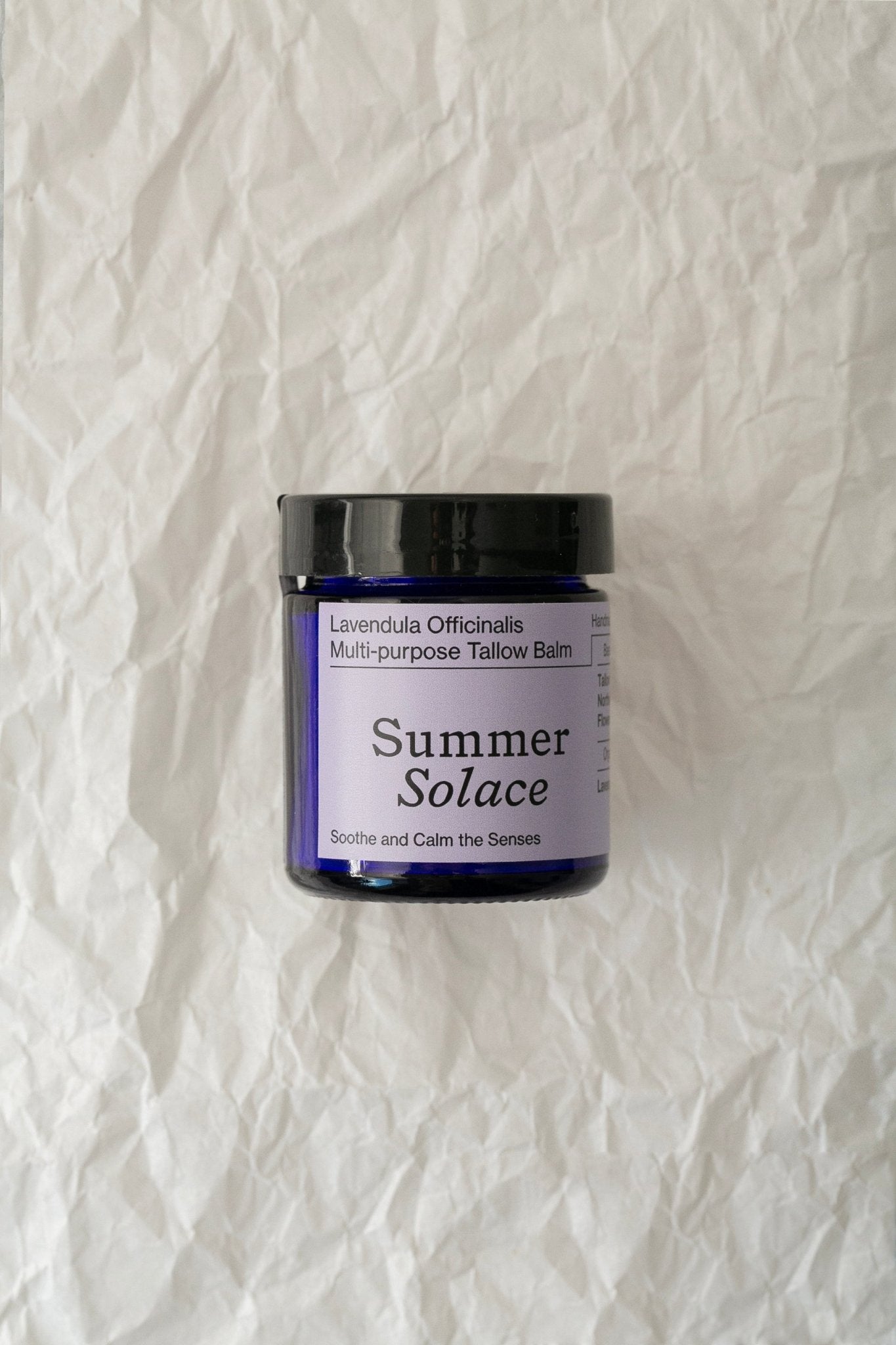 A small jar of "Summer Solace Tallow" Lavendula Officinalis Balm – Regenerative Tallow™ rests against a crinkled white background. The label reads "Lavendula Officinalis," and the balm is intended to soothe, calm the senses, and provide a luxurious moisturizing experience. The jar is a deep blue with a black lid.