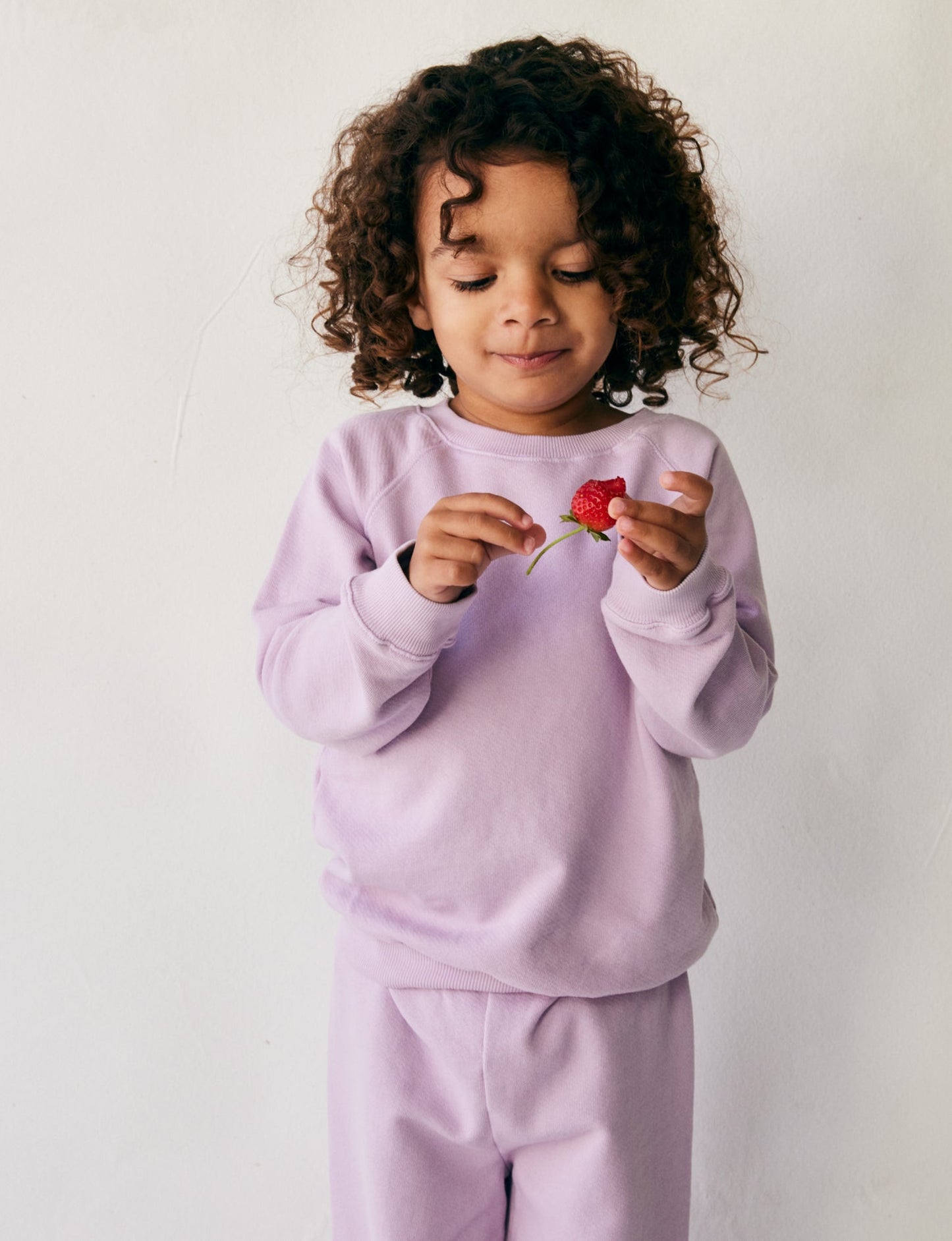 A young child with curly hair stands against a plain white background, wearing The Lil' Classic Crewneck in a vintage-inspired lavender color from the brand all the babies, along with matching pants. They are looking down and smiling softly while holding a small, red flower in their hands.