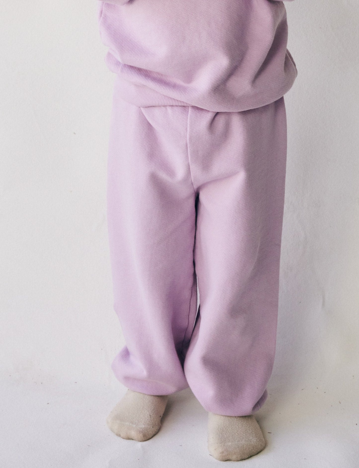 A child in The Lil' Classic Sweatpant and a matching sweatshirt by all the babies, made from organic French terry cloth, is standing with their feet slightly apart. The child is barefoot, and the background is plain white.