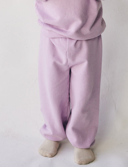 A child in The Lil' Classic Sweatpant and a matching sweatshirt by all the babies, made from organic French terry cloth, is standing with their feet slightly apart. The child is barefoot, and the background is plain white.