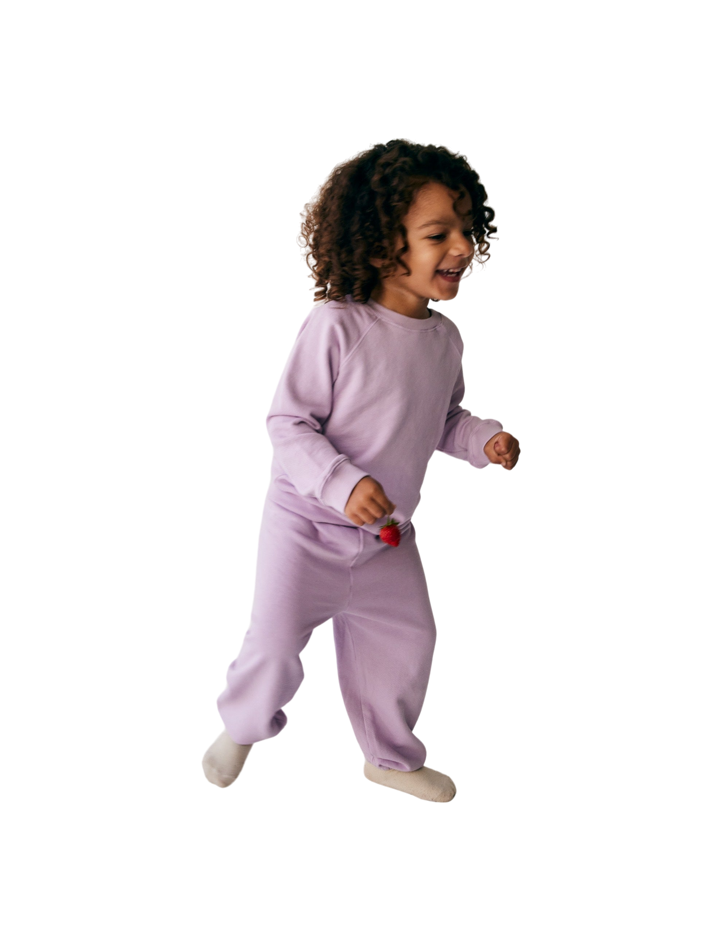 A young child with curly hair is joyfully running, wearing all the babies' The Lil' Classic Crewneck in light purple along with matching pants, both made from organic French terry cloth. Holding a small red object in one hand, the child appears to be indoors and is smiling.