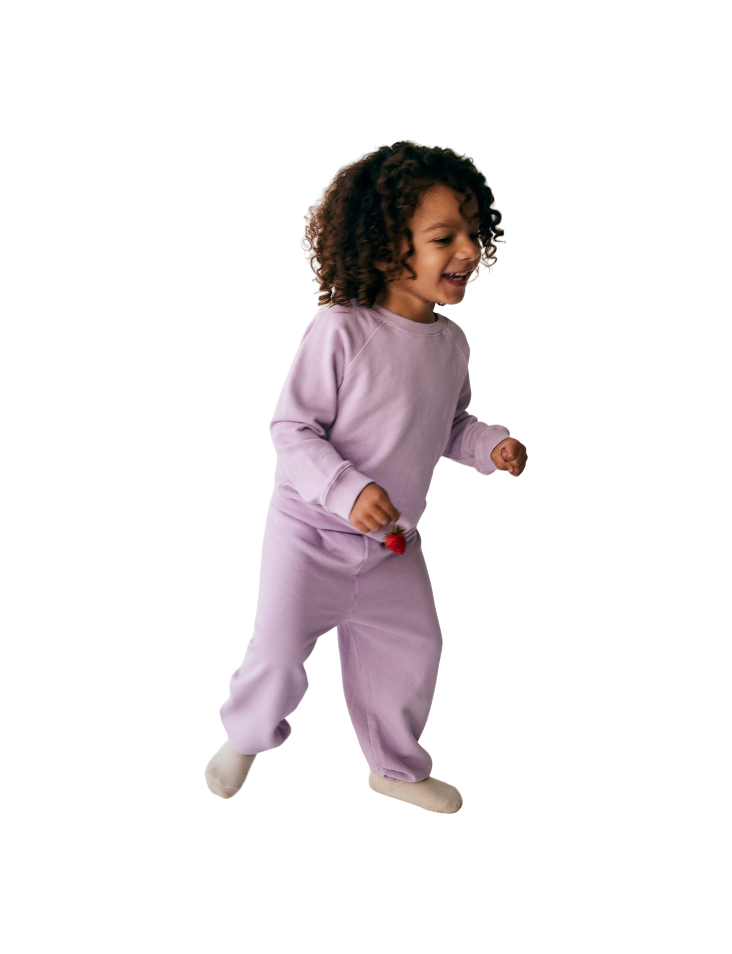 A young child with curly hair is joyfully running, wearing all the babies' The Lil' Classic Crewneck in light purple along with matching pants, both made from organic French terry cloth. Holding a small red object in one hand, the child appears to be indoors and is smiling.