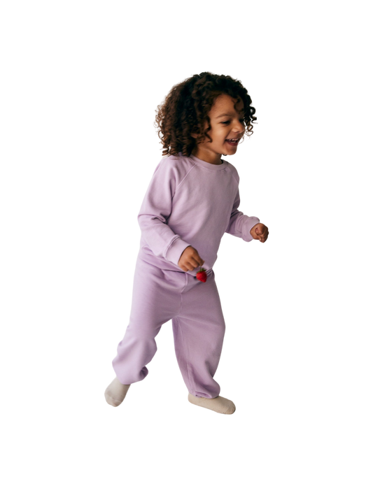 A young child with curly hair is joyfully running, wearing all the babies' The Lil' Classic Crewneck in light purple along with matching pants, both made from organic French terry cloth. Holding a small red object in one hand, the child appears to be indoors and is smiling.