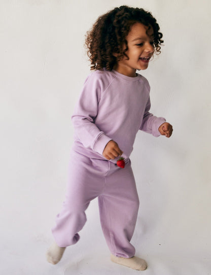 A joyful child with curly hair, donning a cozy closet staple—The Lil' Classic Crewneck in purple made from organic French terry cloth by all the babies—along with white socks, is playing and smiling. The child holds a small toy and is positioned against a plain white background.