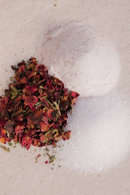 A pile of dried rose petals sits next to two mounds of coarse sea salt on a beige surface. The vibrant red and pink petals contrast with the white, granular salt, creating a visually striking ambiance perfect for Surya's Love Bath Heart Opening Soak.