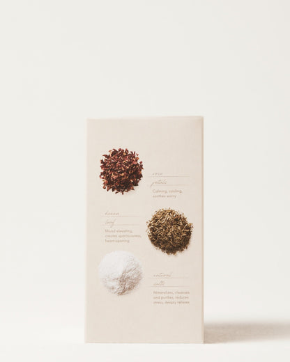 An image of a beige-colored product package for the "Love Bath Heart Opening Soak" by Surya featuring three different dried herbs and supplements. The top section contains a brownish-red herb labeled "rose petals," the middle section has a green herb labeled "lemon balm," and the bottom section contains a white powder labeled "marine minerals." Descriptions accompany each item.