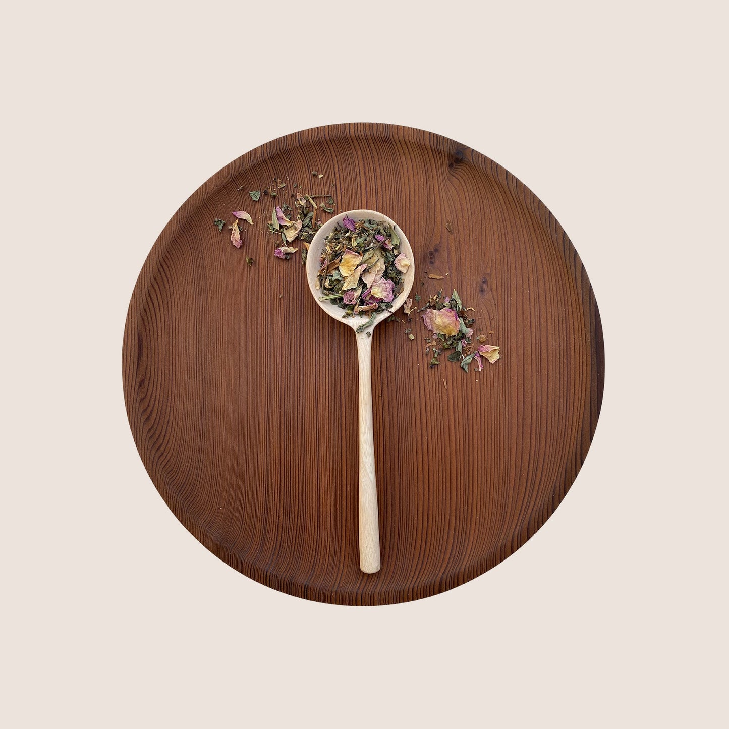 A wooden spoon filled with dried herbs and petals from the Holy Heart Tea Blend by Moon Canyon Healing rests on a round wooden plate. Scattered herbs and petals surround the spoon, creating a simple and rustic composition against a light beige background, evoking heart-centered awareness with adaptogenic ingredients.