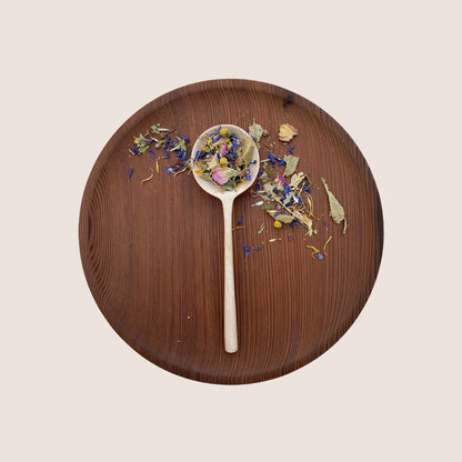 A wooden plate holds a wooden spoon filled with Moon Canyon Healing's Meadow Medicine Tea Blend, a vibrant mix of colorful dry flowers and herbs. Additional dry flower petals and herbs from this inviting herbal blend are scattered around the spoon on the plate. The light beige background highlights the natural colors of these organic tea elements.