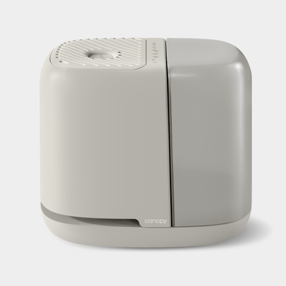 The Bedside Humidifier 2.0 by Canopy is a minimalist, modern light gray appliance with a sleek square design, featuring a textured top and central vent. The brand name is elegantly displayed on the lower front against a plain white background.