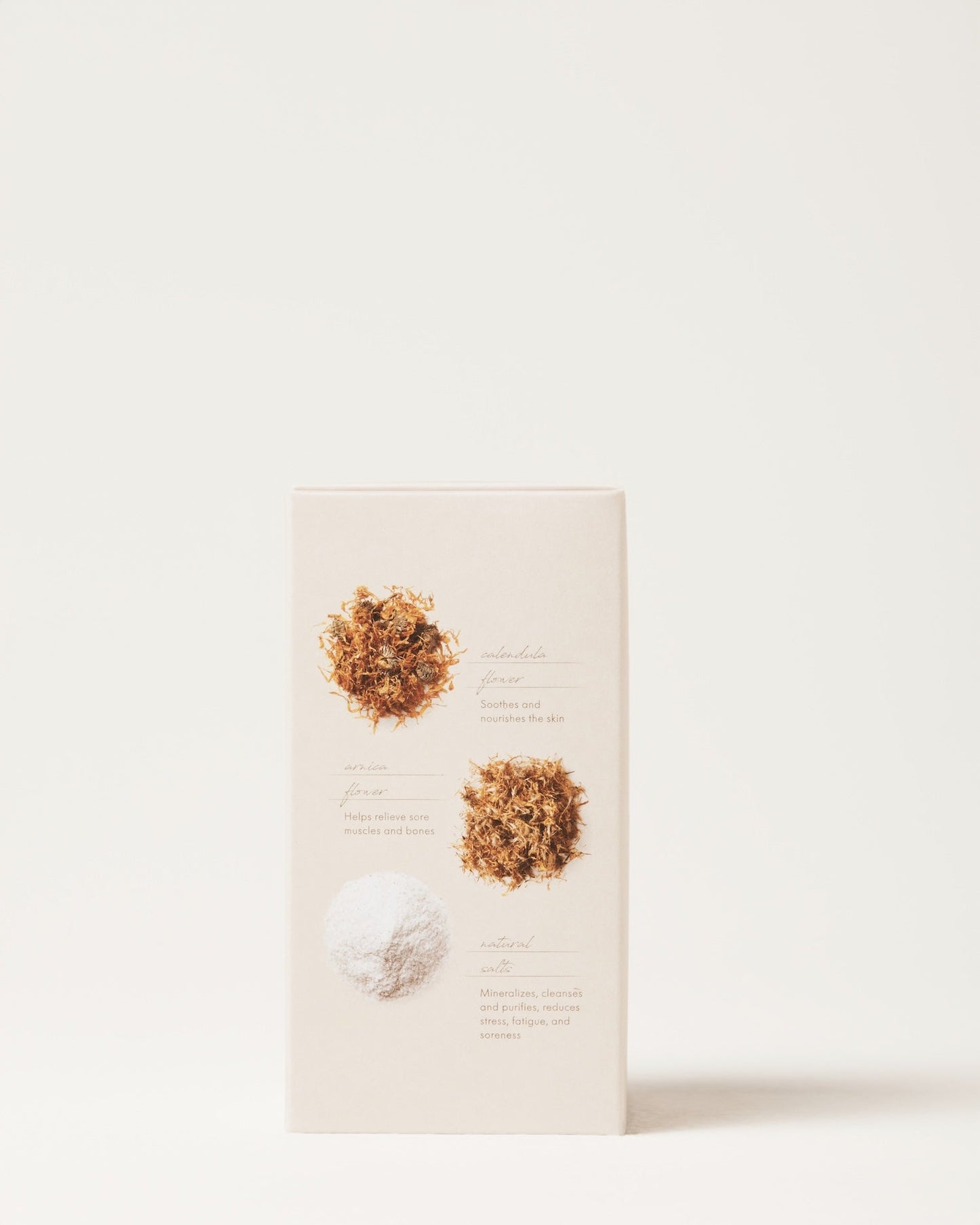 A close-up of the rectangular, beige packaging of the Muscle Relief Bath Soak by Surya against a plain white background. The front of the packaging showcases text and images of three ingredients: two kinds of dried flowers and a white powder, each accompanied by descriptions that highlight its use as an Ayurvedic bath soak for spa therapy.