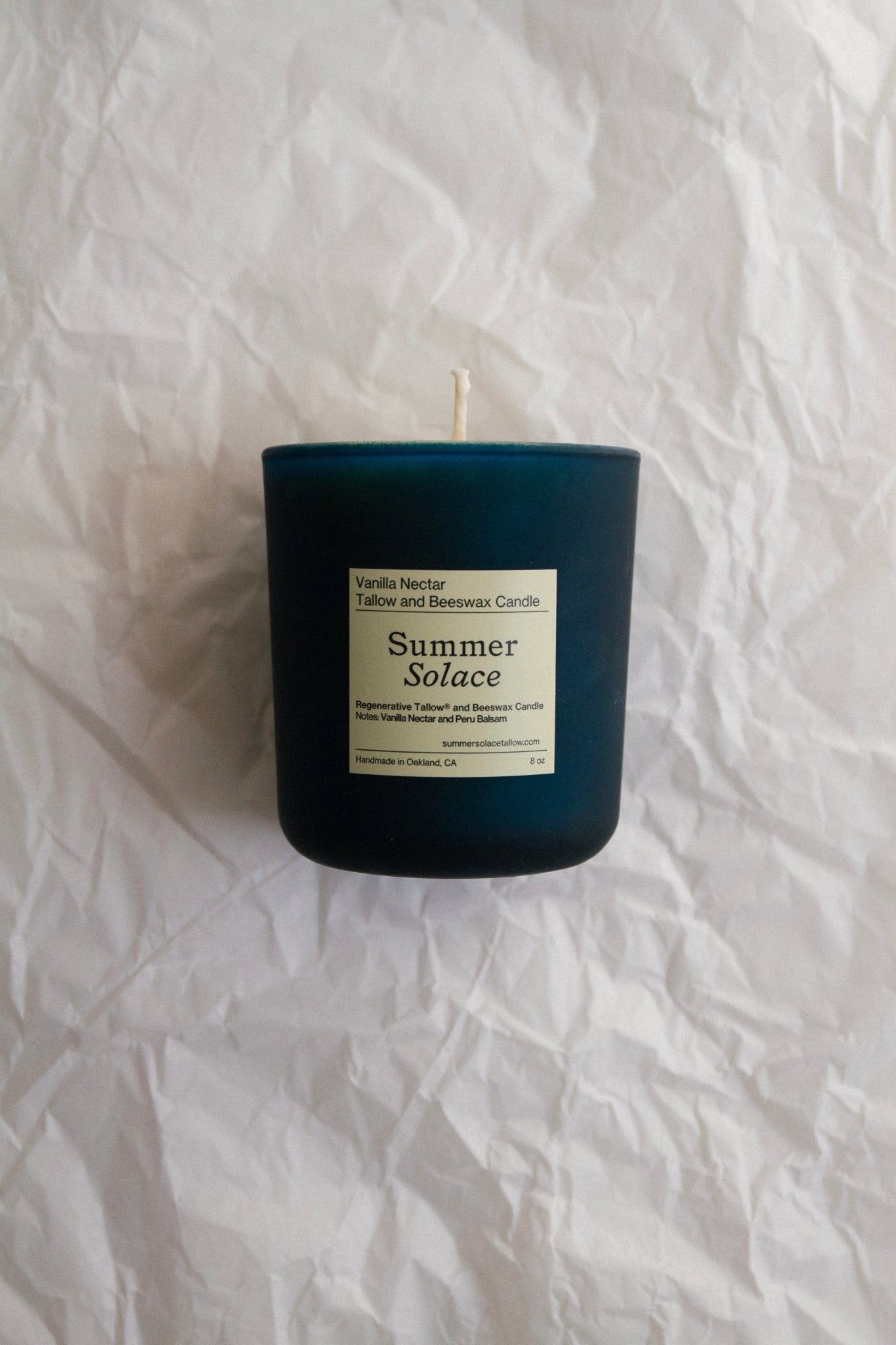 A dark blue Vanilla Nectar + Peru Balsam Tallow and Beeswax Candle (8 oz.) from Summer Solace Tallow, with a label that reads "Summer Solace" and "Vanilla Nectar + Peru Balsam Tallow and Beeswax Candle," is placed on crumpled white paper. The candle’s wick is unlit, and the packaging exudes a natural and handcrafted impression.