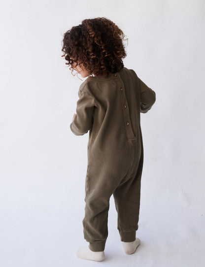 A young child with curly hair is standing and facing away from the camera, wearing The Thermal Onesie by all the babies. The olive-green, long-sleeve onesie features buttons down the back. The child is standing on a white surface in front of a white background.