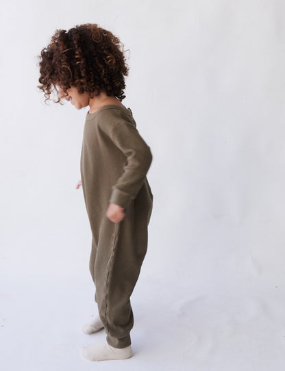 A young child with curly hair stands in profile, wearing The Thermal Onesie by all the babies, a green long-sleeved thermal onesie, paired with light-colored socks. The background is a plain, light-colored wall.