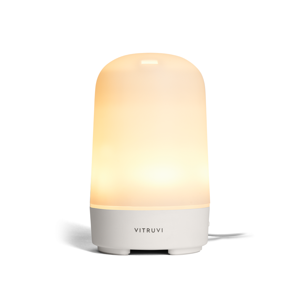 The Vitruvi Glow Diffuser is a sleek, modern essential oil diffuser with a smooth, cylindrical design. It features a white finish and emits a soft, warm light from the top half to create a calming ambiance. Use it with 100% pure essential oils for a serene experience. A cord extends visibly from the back.
