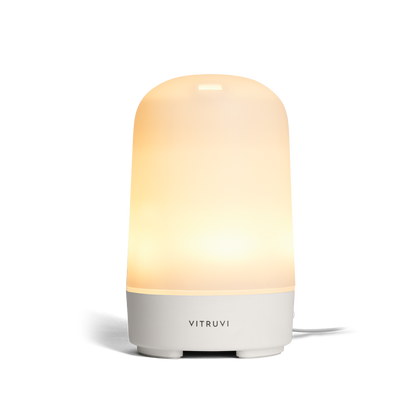 The Vitruvi Glow Diffuser is a sleek, modern essential oil diffuser with a smooth, cylindrical design. It features a white finish and emits a soft, warm light from the top half to create a calming ambiance. Use it with 100% pure essential oils for a serene experience. A cord extends visibly from the back.
