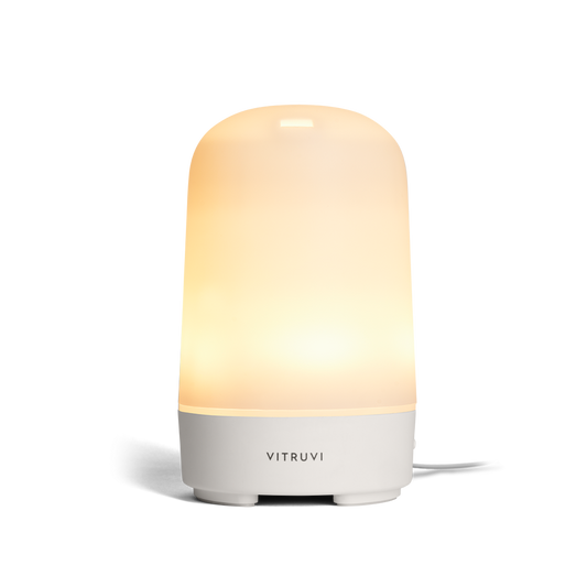 The Vitruvi Glow Diffuser is a sleek, modern essential oil diffuser with a smooth, cylindrical design. It features a white finish and emits a soft, warm light from the top half to create a calming ambiance. Use it with 100% pure essential oils for a serene experience. A cord extends visibly from the back.
