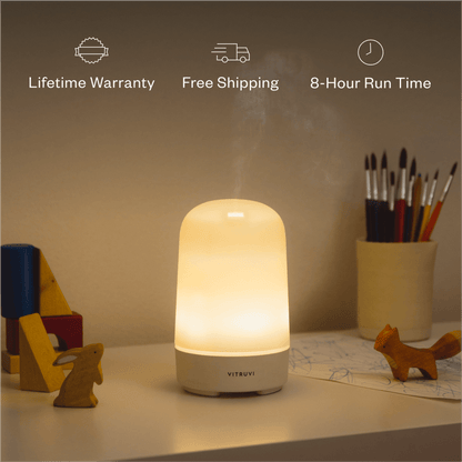 A lit Glow Diffuser by Vitruvi sits on a table surrounded by small wooden animal figures and art supplies in a dimly lit room. Text above the Glow Diffuser highlights "Lifetime Warranty," "Free Shipping," and "8-Hour Run Time.