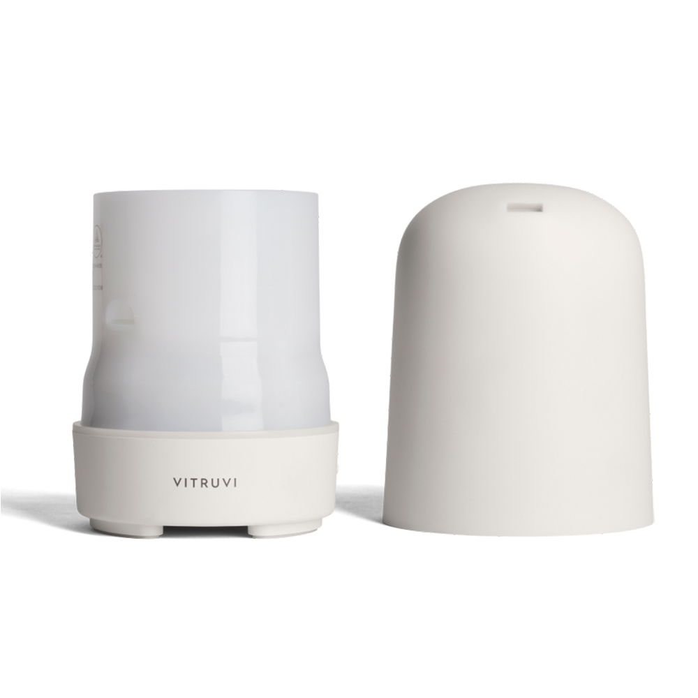 A white Glow Diffuser by Vitruvi with its cover removed is displayed. The sleek, cylindrical design features a smooth base with the brand name "Vitruvi" on it. The cover is placed next to the diffuser, revealing the inner chamber for adding 100% pure essential oils and water.