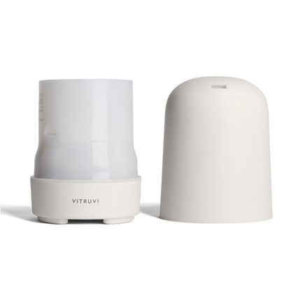 A white Glow Diffuser by Vitruvi with its cover removed is displayed. The sleek, cylindrical design features a smooth base with the brand name "Vitruvi" on it. The cover is placed next to the diffuser, revealing the inner chamber for adding 100% pure essential oils and water.