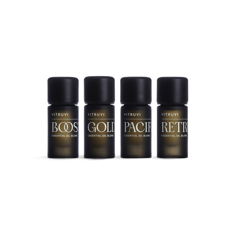 Four dark brown glass bottles with black caps feature a variety of essential oil blends by Vitruvi. The labels read: BOOST, GOLD, PACIF, and RETR. This non-toxic Refresh Essential Oil Kit is perfect for creating a serene environment. The white background highlights the beautifully designed Vitruvi products.