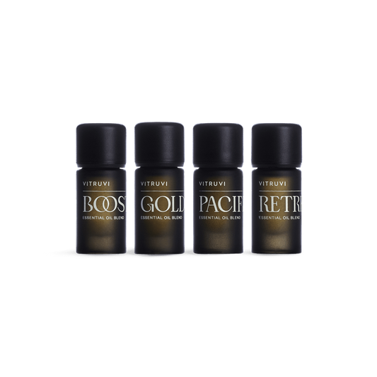 Four dark brown glass bottles with black caps feature a variety of essential oil blends by Vitruvi. The labels read: BOOST, GOLD, PACIF, and RETR. This non-toxic Refresh Essential Oil Kit is perfect for creating a serene environment. The white background highlights the beautifully designed Vitruvi products.