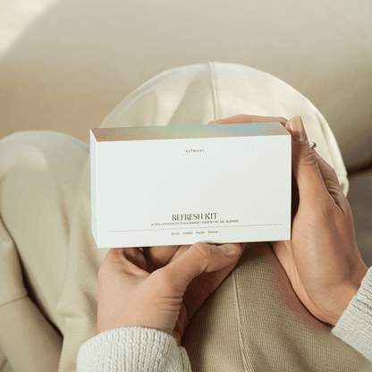 A person dressed in light-colored pants and a sweater holds a white box labeled "Vitruvi Refresh Diffuser Blend Kit," showcasing four premium essential oil blends: Boost, Golden, Pacific, and Retreat. These invigorating aromas are designed to uplift your senses.